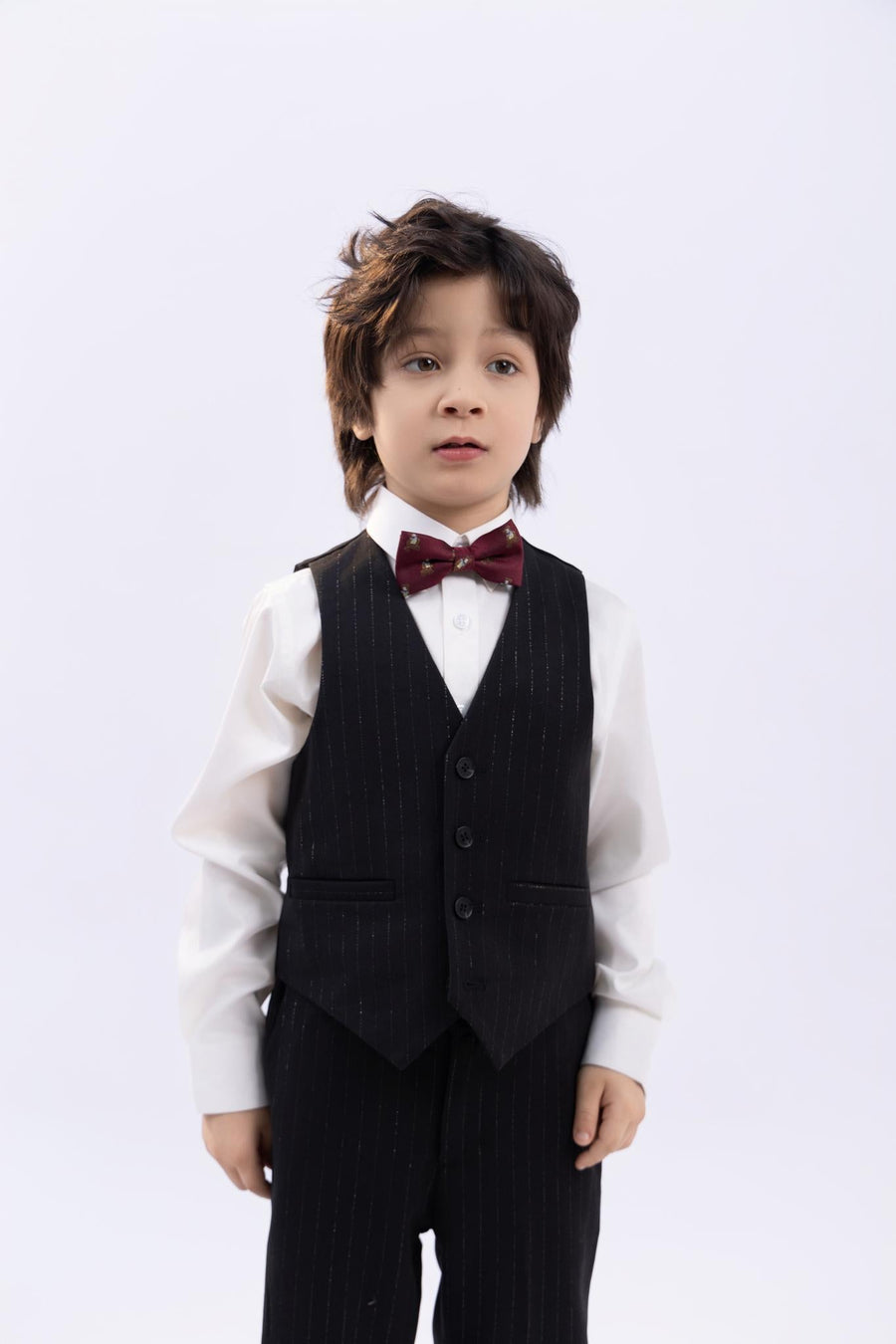 A young boy with tousled hair wears a white shirt, black pinstripe vest and pants, and a KIKI and JOJO Kids Bow Tie – Burgundy Bear (pre-tied satin, adjustable), standing against a plain light background.