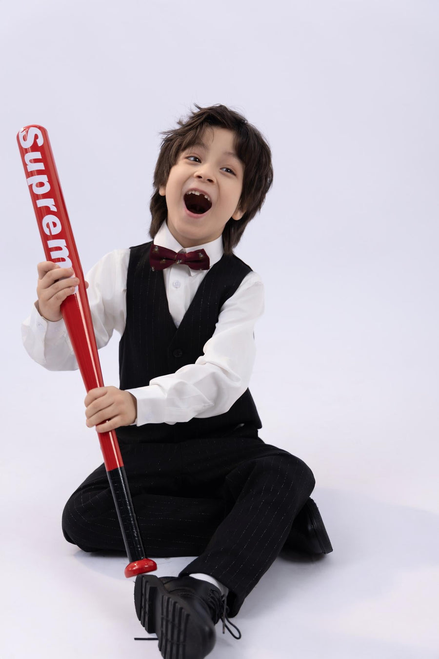 A young boy in a suit and a KIKI and JOJO Kids Bow Tie – Burgundy Bear – Pre-Tied Satin – Adjustable sits on the floor, smiling widely while holding a red baseball bat with "Supreme" printed on it.