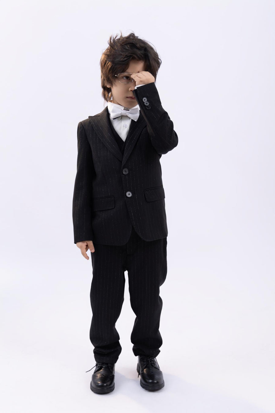 A young boy wearing a KIKI and JOJO Kids Bow Tie – White & Black – Pre-Tied Satin – Adjustable stands against a plain background, covering his eyes with one hand.