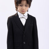 A young child with dark hair and glasses, wearing a classic black and white pinstripe suit, white dress shirt, and a KIKI and JOJO Kids Bow Tie – White & Black – Pre-Tied Satin – Adjustable, stands against a plain light background.