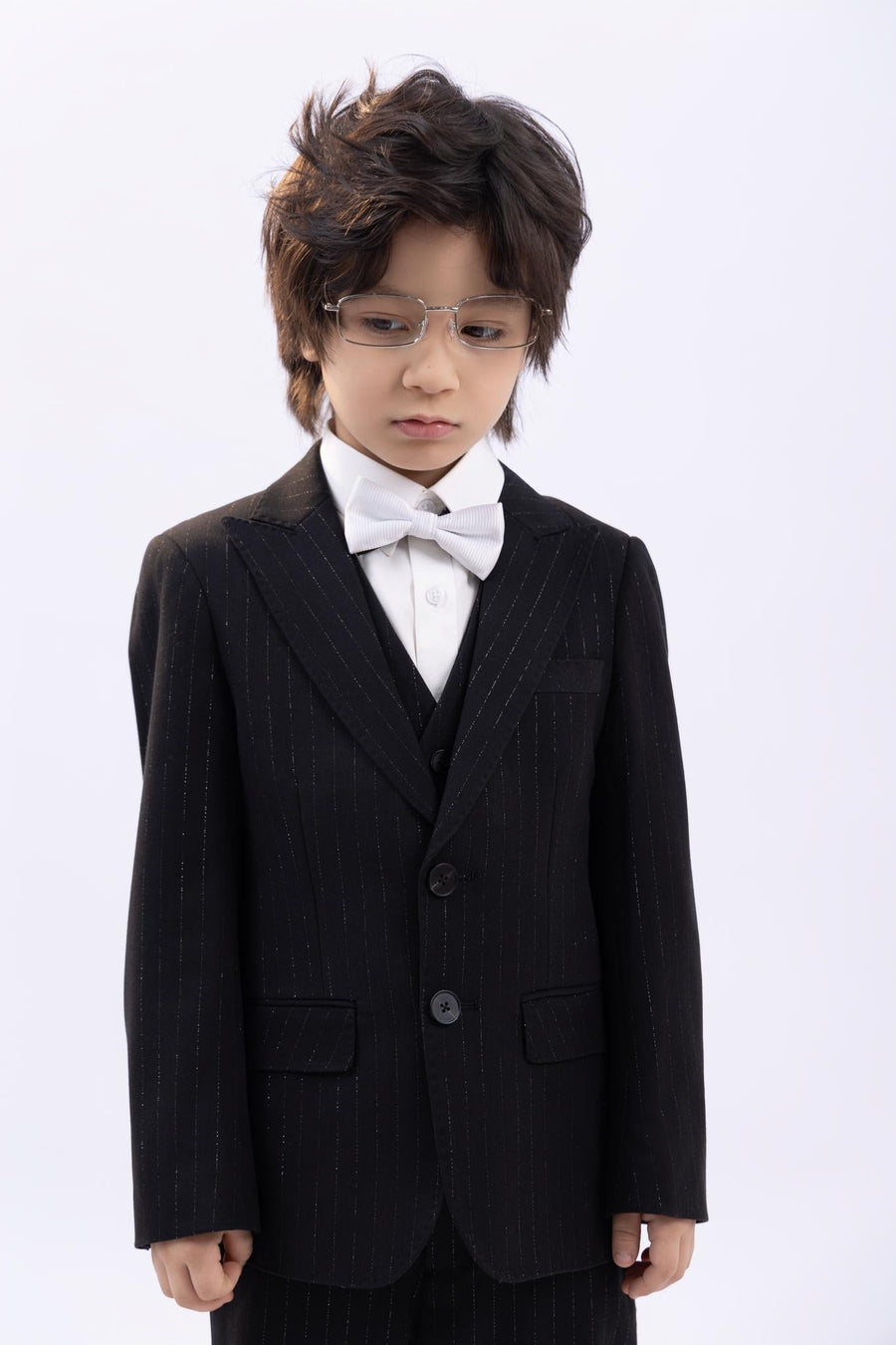 A young child with dark hair and glasses, wearing a classic black and white pinstripe suit, white dress shirt, and a KIKI and JOJO Kids Bow Tie – White & Black – Pre-Tied Satin – Adjustable, stands against a plain light background.