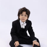 A young boy in a KIKI and JOJO Kids Bow Tie – White & Black – Pre-Tied Satin (adjustable), black suit, and glasses sits against a plain white background.