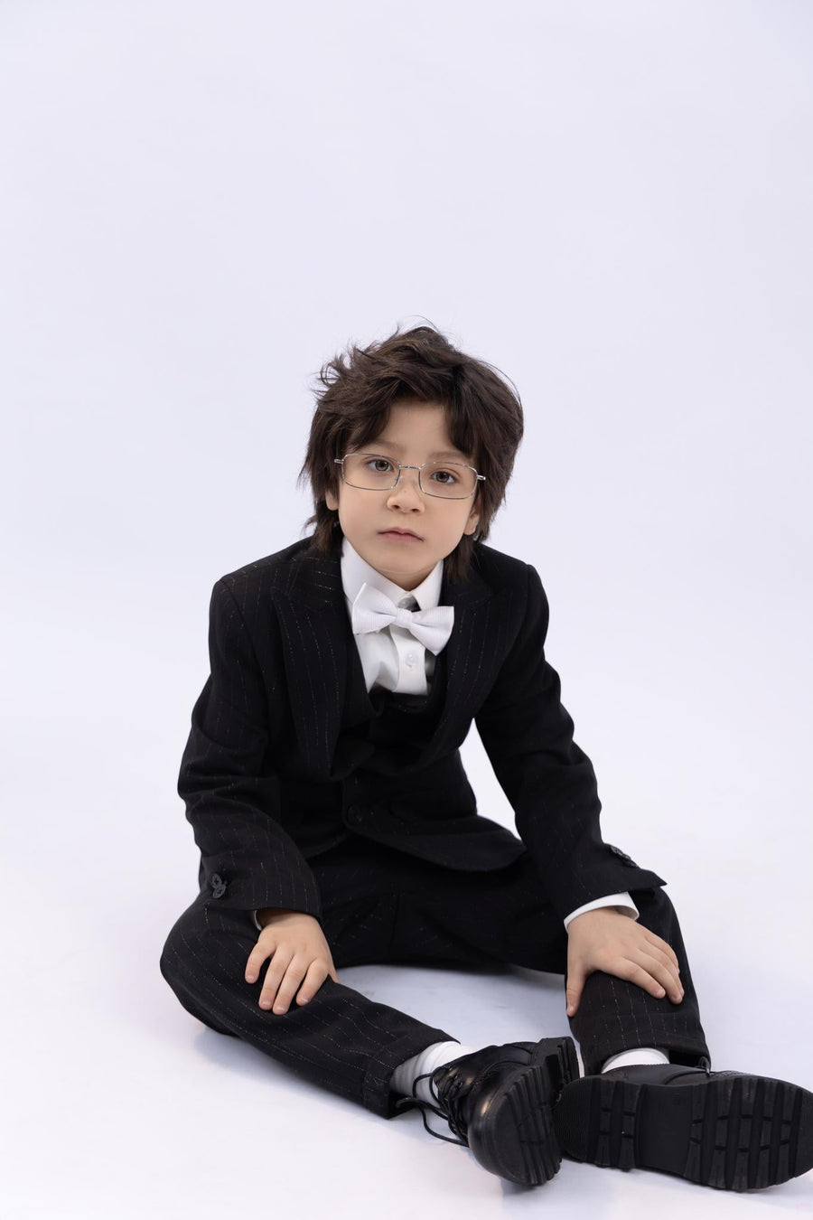 A young boy in a KIKI and JOJO Kids Bow Tie – White & Black – Pre-Tied Satin (adjustable), black suit, and glasses sits against a plain white background.