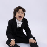 A young boy wearing the KIKI and JOJO Kids Bow Tie – White & Black – Pre-Tied Satin, with glasses and black shoes, sits on the floor against a plain white background, his mouth open in surprise.
