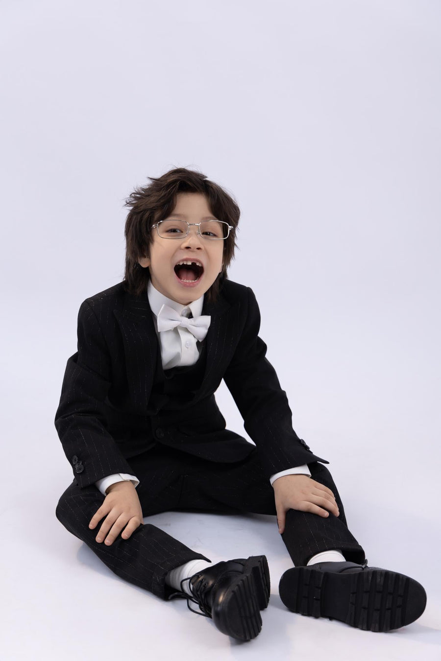 A young boy wearing the KIKI and JOJO Kids Bow Tie – White & Black – Pre-Tied Satin, with glasses and black shoes, sits on the floor against a plain white background, his mouth open in surprise.
