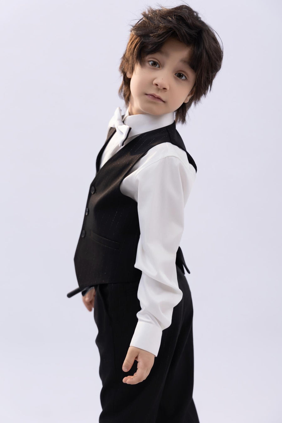 A young boy from KIKI and JOJO wears the Kids Bow Tie – White & Black – Pre-Tied Satin – Adjustable, standing slightly turned and looking at the camera against a plain light background.