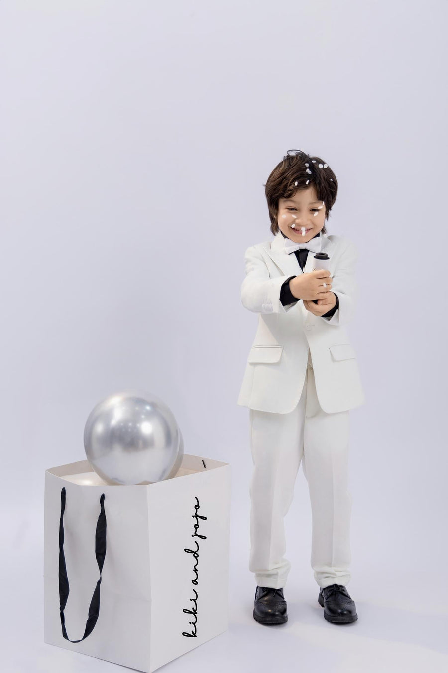 A young boy in a classic KIKI and JOJO Kids Bow Tie – White & Black – Pre-Tied Satin with adjustable fit smiles while popping a party popper, celebrating in style next to a gift bag with a silver balloon inside.