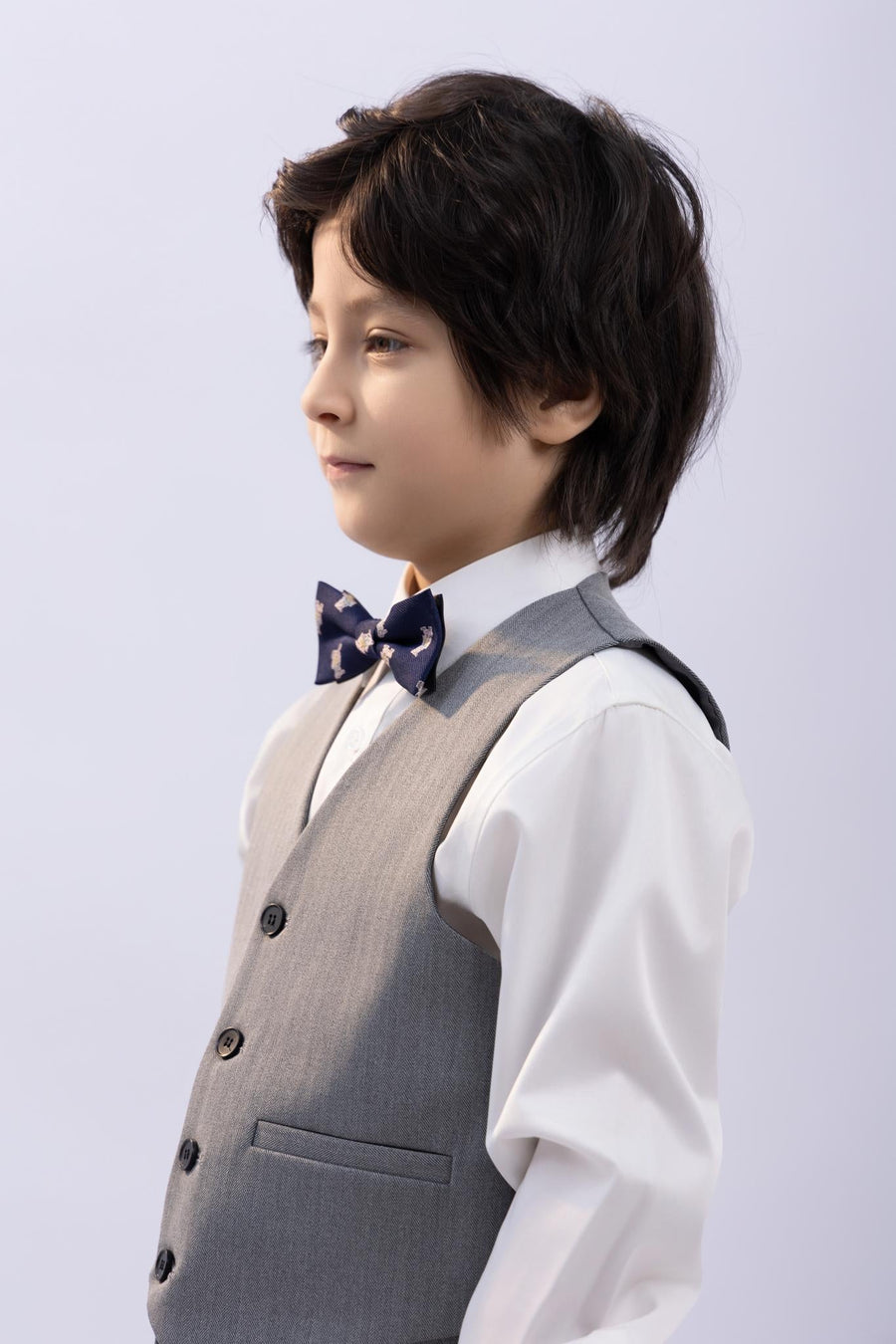 A young boy with dark hair wears a white shirt, grey vest, and the KIKI and JOJO Kids Bow Tie – Navy Blue Bear – Pre-Tied Satin – Adjustable, standing in profile against a plain light background.