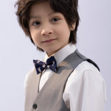 A young boy with dark hair, in a white shirt and gray vest, wears the KIKI and JOJO Kids Bow Tie – Navy Blue Bear – Pre-Tied Satin – Adjustable, standing in front of a plain light background.