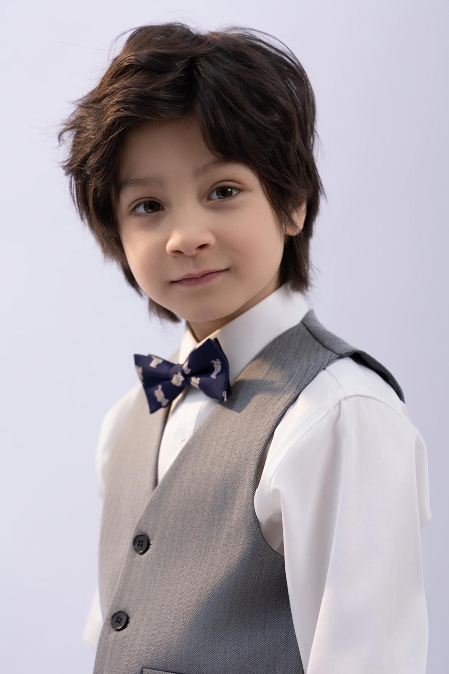 A young boy with dark hair, in a white shirt and gray vest, wears the KIKI and JOJO Kids Bow Tie – Navy Blue Bear – Pre-Tied Satin – Adjustable, standing in front of a plain light background.
