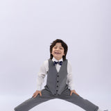 A young boy in a gray suit, white shirt, and KIKI and JOJO's Navy Blue Bear Pre-Tied Satin Kids Bow Tie poses in a wide-legged split, smiling at the camera against a plain light background.