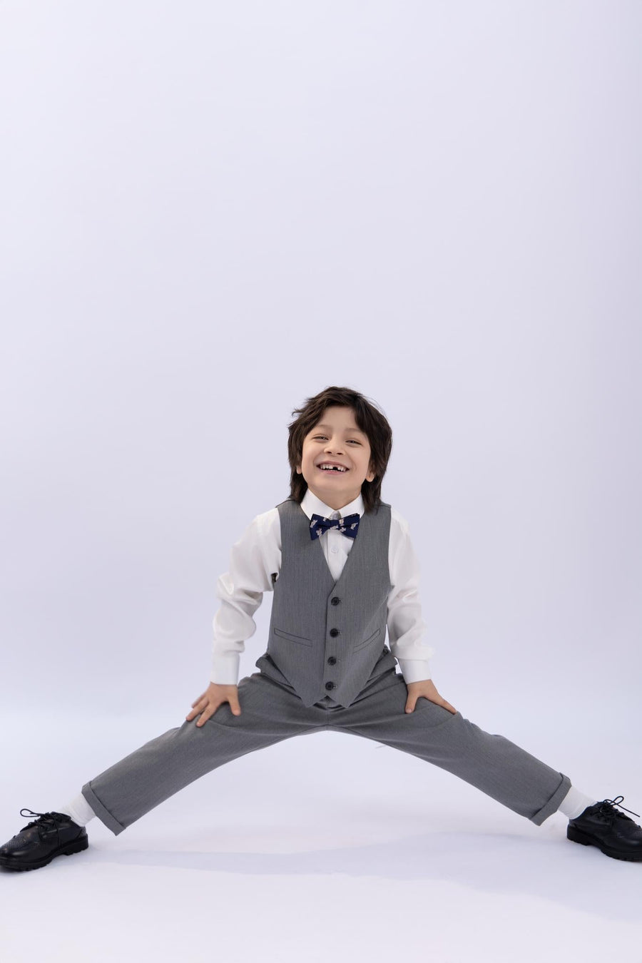 A young boy in a gray suit, white shirt, and KIKI and JOJO's Navy Blue Bear Pre-Tied Satin Kids Bow Tie poses in a wide-legged split, smiling at the camera against a plain light background.