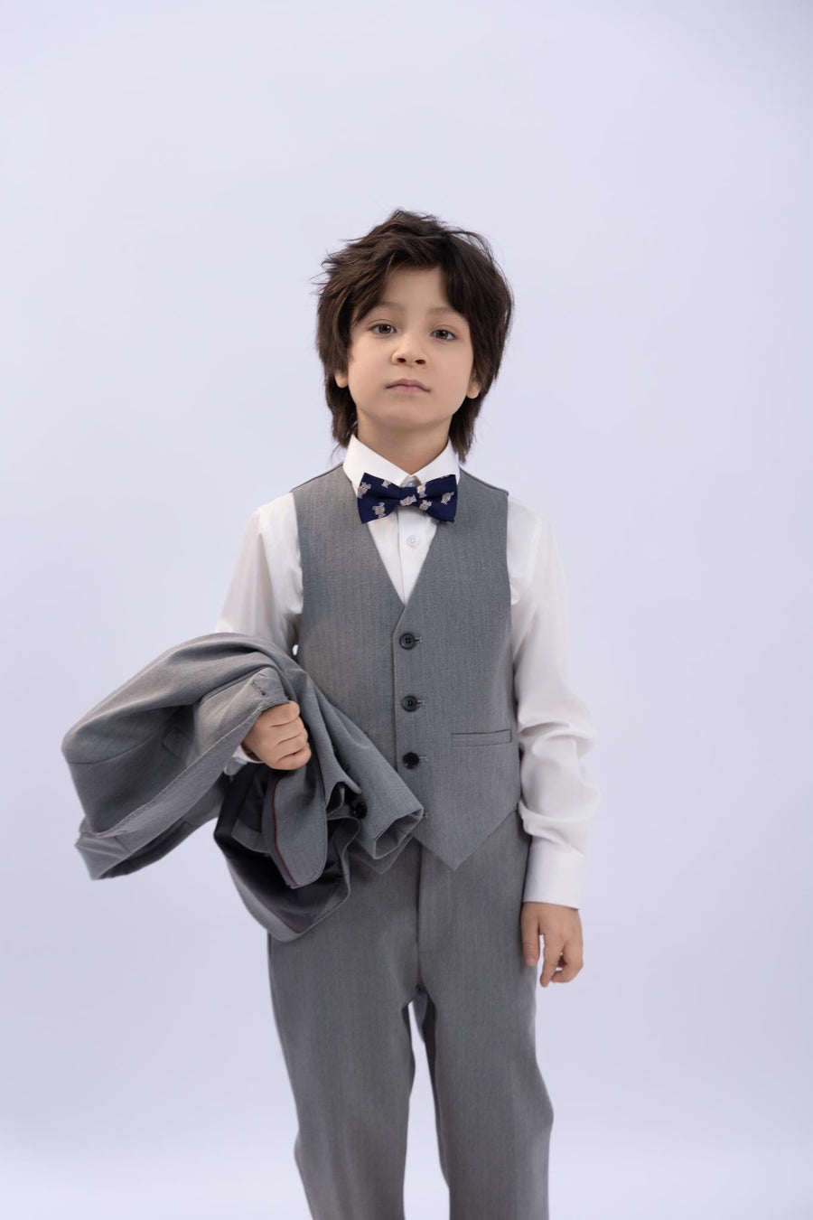 A young boy in a gray suit wears the KIKI and JOJO Kids Bow Tie – Navy Blue Bear – Pre-Tied Satin – Adjustable, standing against a plain background and holding his premium suit jacket in his left hand.