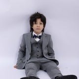 A young boy in a gray suit wears the KIKI and JOJO Kids Bow Tie – Navy Blue Bear – Pre-Tied Satin – Adjustable, sitting on the floor against a plain white background, looking forward with a neutral expression.