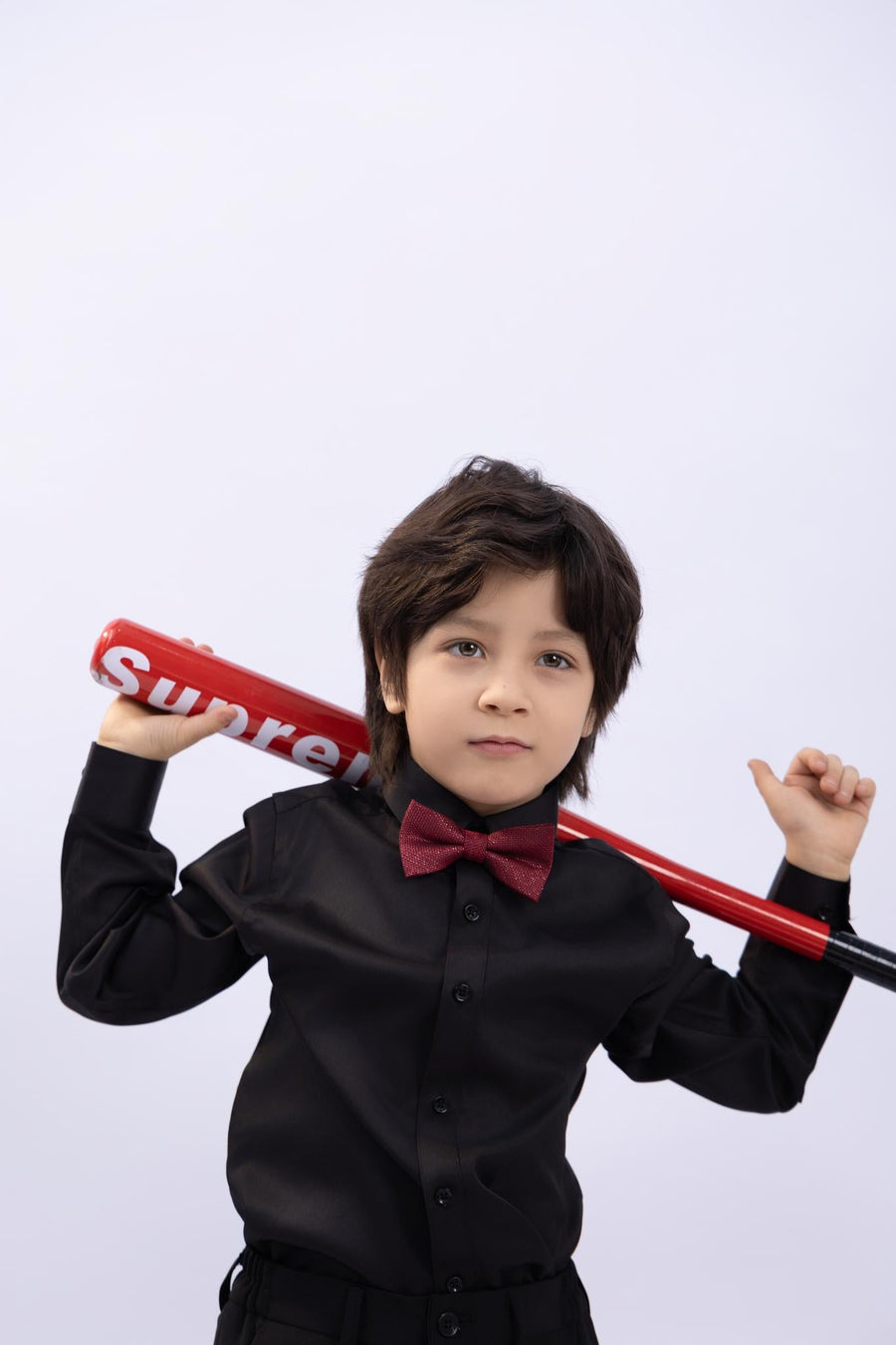 A young boy in a black shirt wears the KIKI and JOJO Kids Bow Tie – Burgundy & Black, a pre-tied adjustable satin accessory, while holding a red baseball bat over his shoulders against a plain light background.