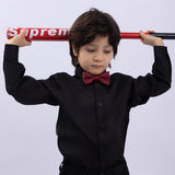 A young boy in a black shirt and KIKI and JOJO Kids Bow Tie – Burgundy & Black holds a red "Supreme" baseball bat behind his shoulders against a plain light background.