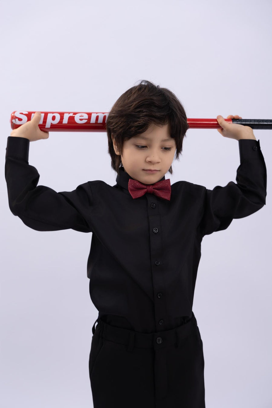 A young boy in a black shirt and KIKI and JOJO Kids Bow Tie – Burgundy & Black holds a red "Supreme" baseball bat behind his shoulders against a plain light background.