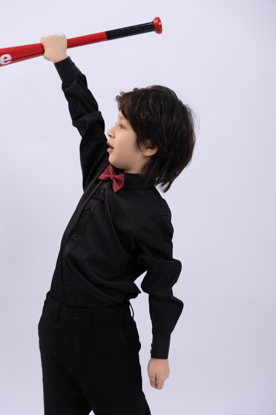 A young boy in a black shirt and KIKI and JOJO’s Kids Bow Tie – Burgundy & Black holds a red baseball bat above his head, highlighting this stylish pre-tied satin bow tie’s premium quality and adjustable fit.