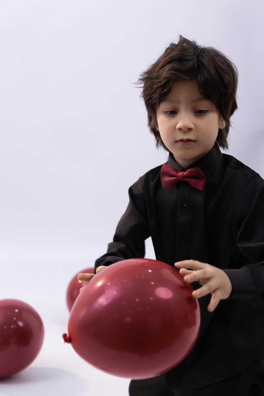 A young boy in a black shirt wears the KIKI and JOJO Kids Bow Tie – Burgundy & Black, a pre-tied satin adjustable bow tie, and holds a red balloon with more red balloons on the floor against a plain background.