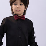 A young boy wears a KIKI and JOJO Kids Bow Tie in Burgundy & Black, pre-tied satin with an adjustable fit, over a black dress shirt, standing against a plain light background and looking slightly to the side.