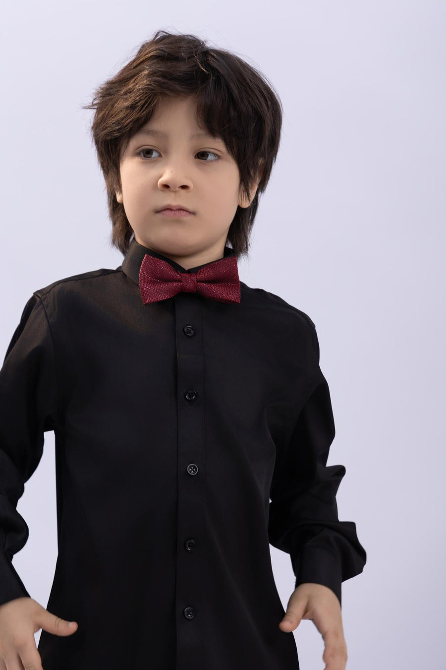 A young boy wears a KIKI and JOJO Kids Bow Tie in Burgundy & Black, pre-tied satin with an adjustable fit, over a black dress shirt, standing against a plain light background and looking slightly to the side.