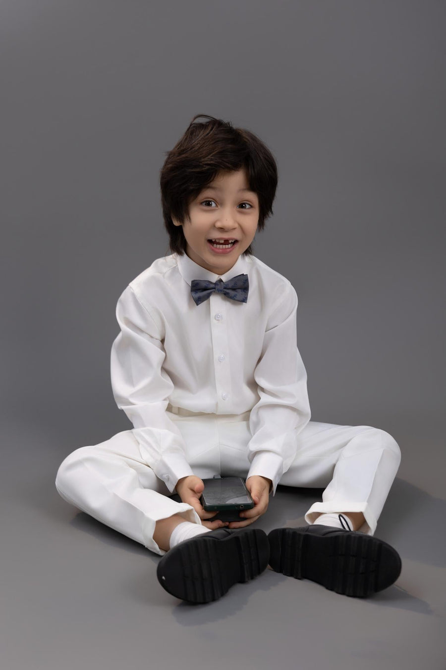 A young boy wearing a KIKI and JOJO Kids Bow Tie – Light Gray Bicycle, a white dress shirt with blue bicycle embroidery, and white pants sits cross-legged on the floor holding a smartphone against a plain gray background.