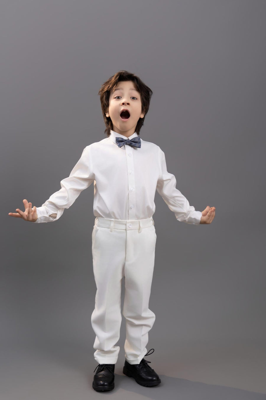 A young boy wearing a crisp white shirt and pants pairs his look with the KIKI and JOJO Kids Bow Tie – Light Gray Bicycle, a pre-tied, adjustable satin accessory, as he stands against a gray background with mouth open and arms extended.