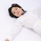 A young boy with dark hair, wearing a KIKI and JOJO Kids Bow Tie – Light Gray Bicycle (pre-tied satin, adjustable) and a white shirt, lies on his back with arms outstretched on a white surface.