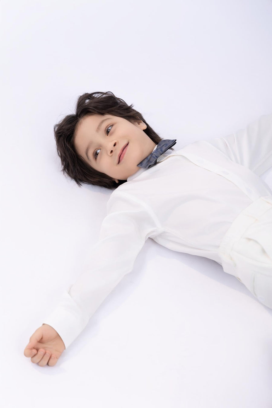 A young boy with dark hair, wearing a KIKI and JOJO Kids Bow Tie – Light Gray Bicycle (pre-tied satin, adjustable) and a white shirt, lies on his back with arms outstretched on a white surface.