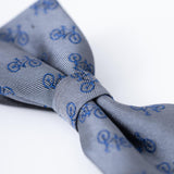 A close-up of the KIKI and JOJO Kids Bow Tie – Light Gray Bicycle, a pre-tied satin bow tie with an adjustable fit, featuring a pattern of small blue bicycles on a light gray background.