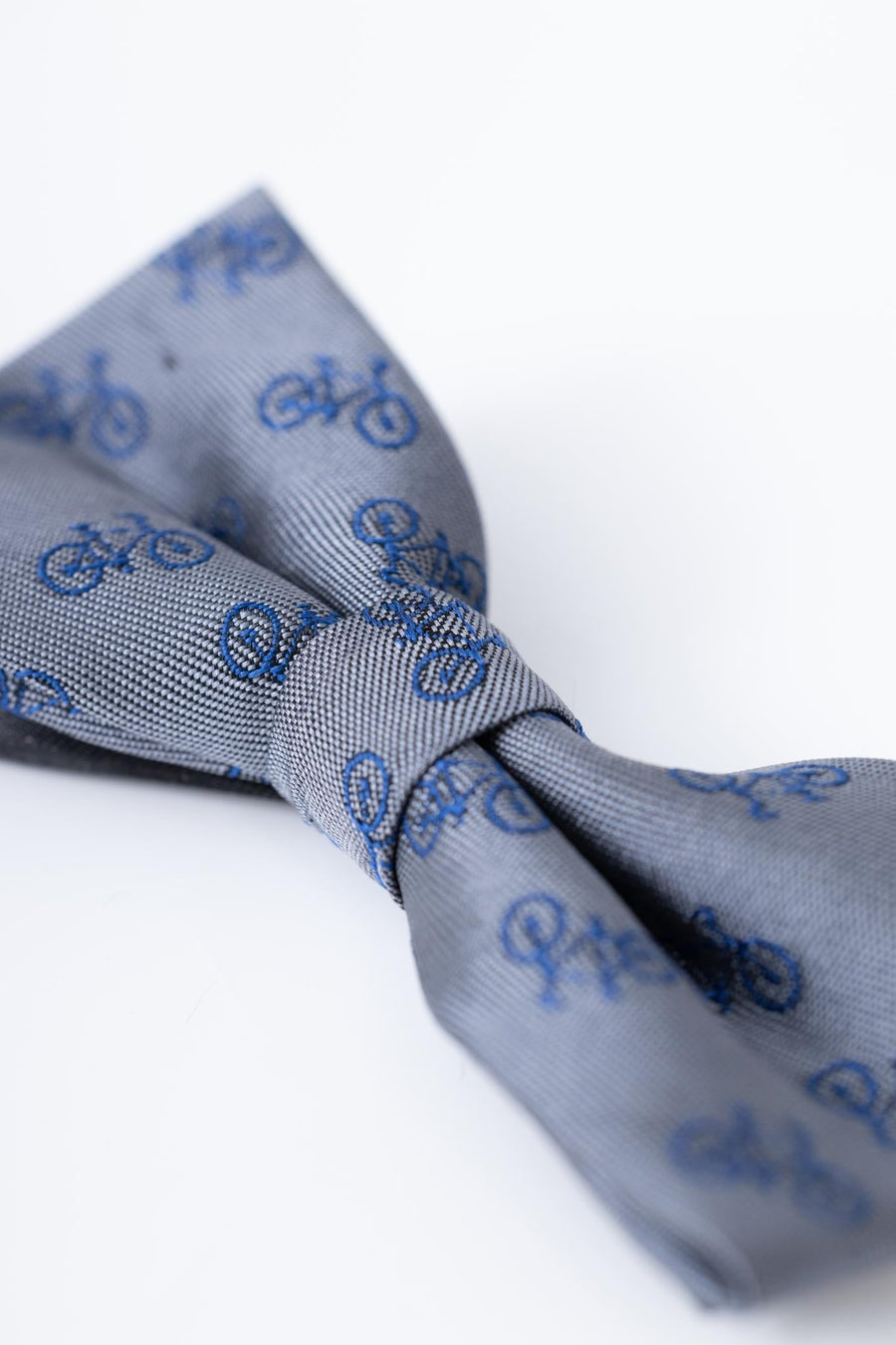 A close-up of the KIKI and JOJO Kids Bow Tie – Light Gray Bicycle, a pre-tied satin bow tie with an adjustable fit, featuring a pattern of small blue bicycles on a light gray background.