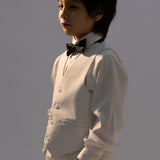 A young boy stands with his hands in his pockets, looking to the side against a plain background, wearing a kids dress shirt, suit, vest, and a KIKI and JOJO Kids Bow Tie—Black with White Dots, pre-tied satin and adjustable.