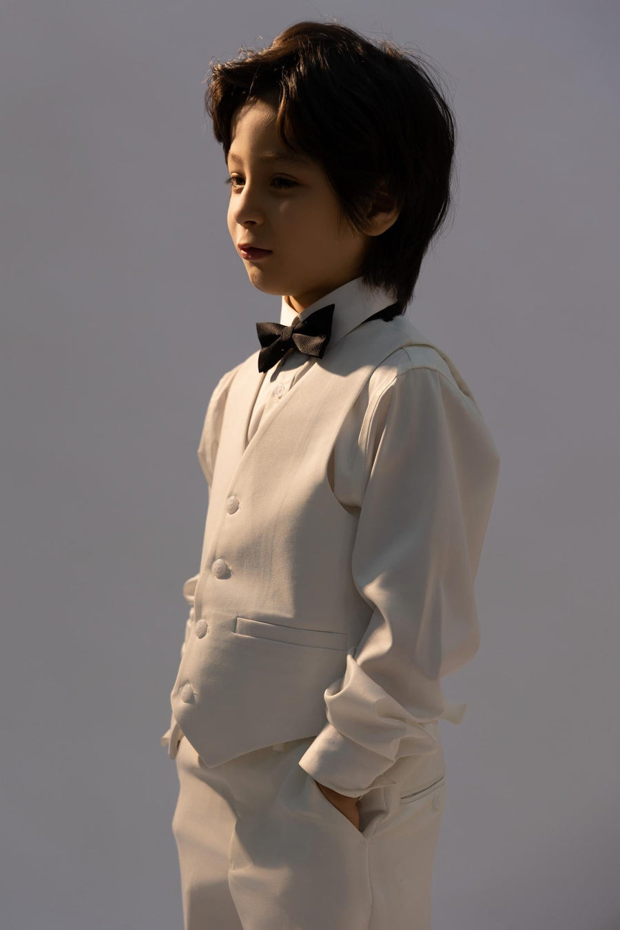 A young boy stands with his hands in his pockets, looking to the side against a plain background, wearing a kids dress shirt, suit, vest, and a KIKI and JOJO Kids Bow Tie—Black with White Dots, pre-tied satin and adjustable.