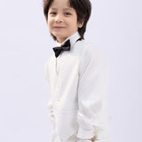 A young boy, dressed in a crisp white suit and premium shirt, smiles at the camera while wearing the KIKI and JOJO Kids Bow Tie – Black with White Dots – Pre-Tied Satin – Adjustable, standing against a plain light background.