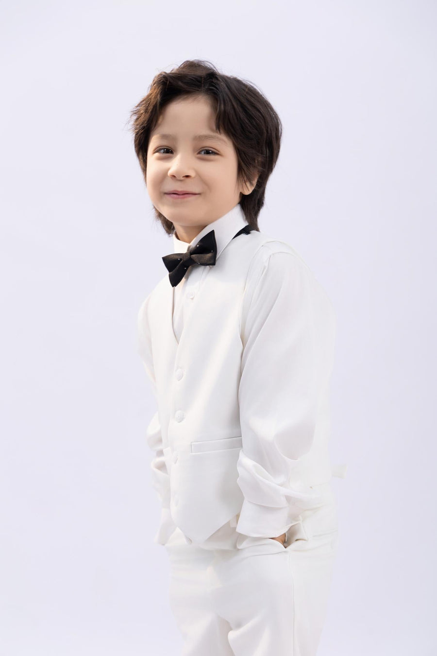 A young boy, dressed in a crisp white suit and premium shirt, smiles at the camera while wearing the KIKI and JOJO Kids Bow Tie – Black with White Dots – Pre-Tied Satin – Adjustable, standing against a plain light background.
