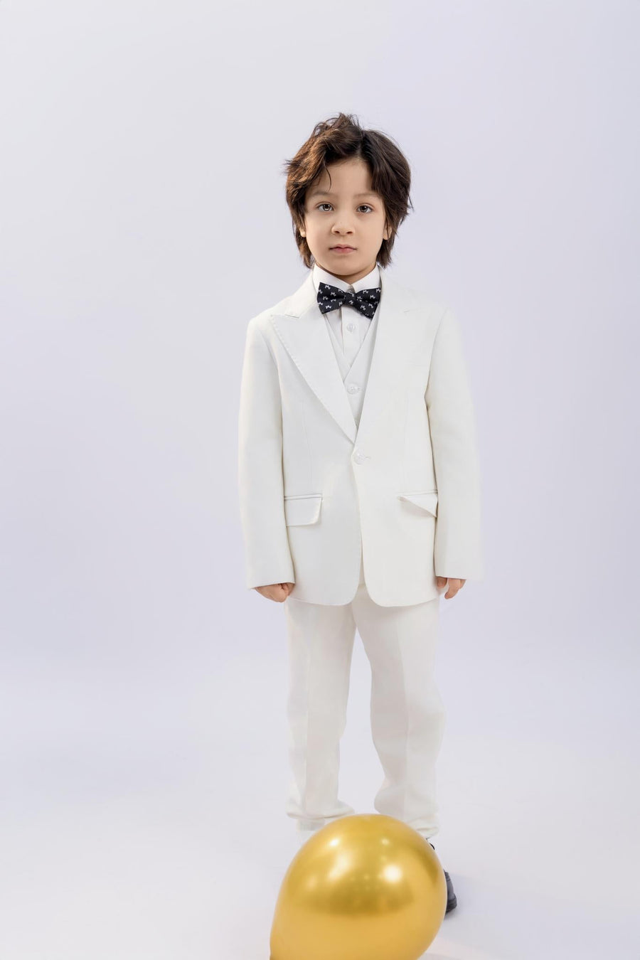 A young boy in a white suit wears the KIKI and JOJO Kids Bow Tie – Black Puppy, a pre-tied satin adjustable bow tie, as he stands on a light background with a gold balloon at his feet.