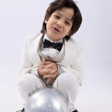 A young boy in a white suit smiles while kneeling with hands clasped, wearing the KIKI and JOJO Kids Bow Tie – Black Puppy – Pre-Tied Satin – Adjustable, with a silver balloon in front of him against a light background.