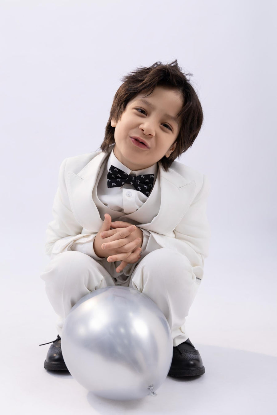 A young boy in a white suit smiles while kneeling with hands clasped, wearing the KIKI and JOJO Kids Bow Tie – Black Puppy – Pre-Tied Satin – Adjustable, with a silver balloon in front of him against a light background.