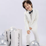 A young boy wears the KIKI and JOJO Kids Bow Tie – Black Puppy – Pre-Tied Satin, standing happily beside a white gift bag and silver balloons against a plain light background.