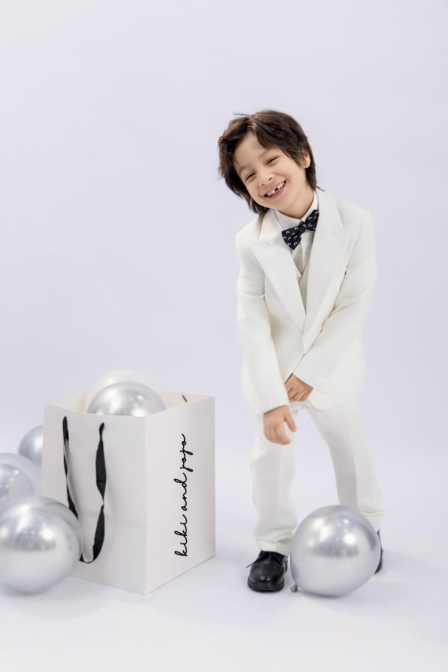 A young boy wears the KIKI and JOJO Kids Bow Tie – Black Puppy – Pre-Tied Satin, standing happily beside a white gift bag and silver balloons against a plain light background.