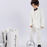 A young boy in a white suit wears the KIKI and JOJO Kids Bow Tie – Black Puppy, a pre-tied adjustable satin bow tie, while standing by silver balloons and a white shopping bag against a plain backdrop.