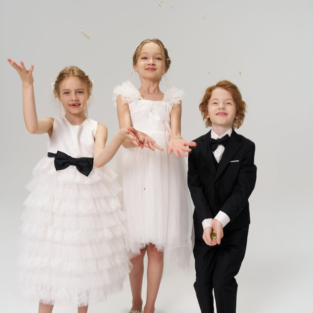 Three children dressed formally pose in a studio; two girls in white dresses toss flower petals, while a boy in a black suit stands holding some petals.