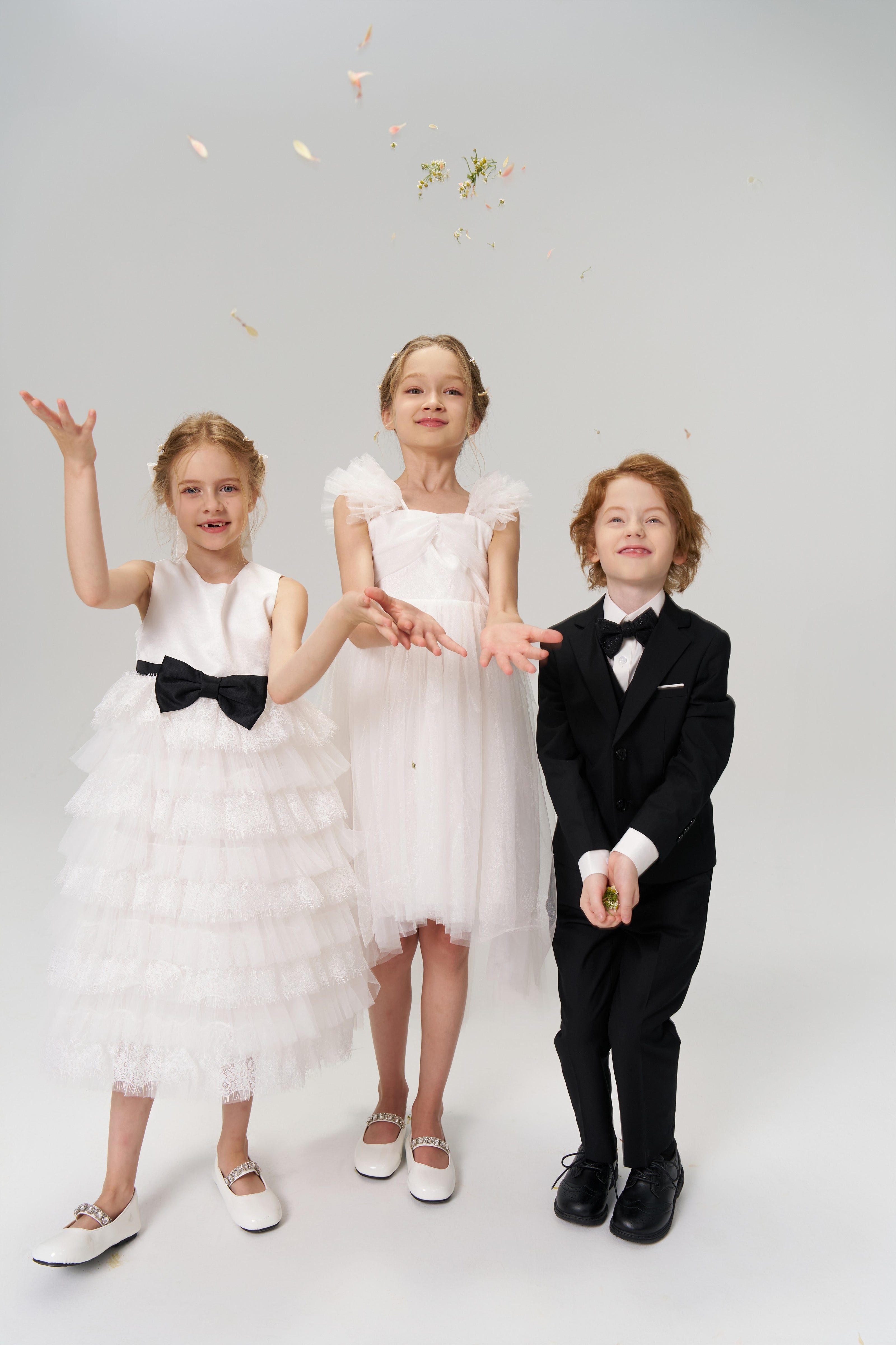 Three children dressed formally pose in a studio; two girls in white dresses toss flower petals, while a boy in a black suit stands holding some petals.