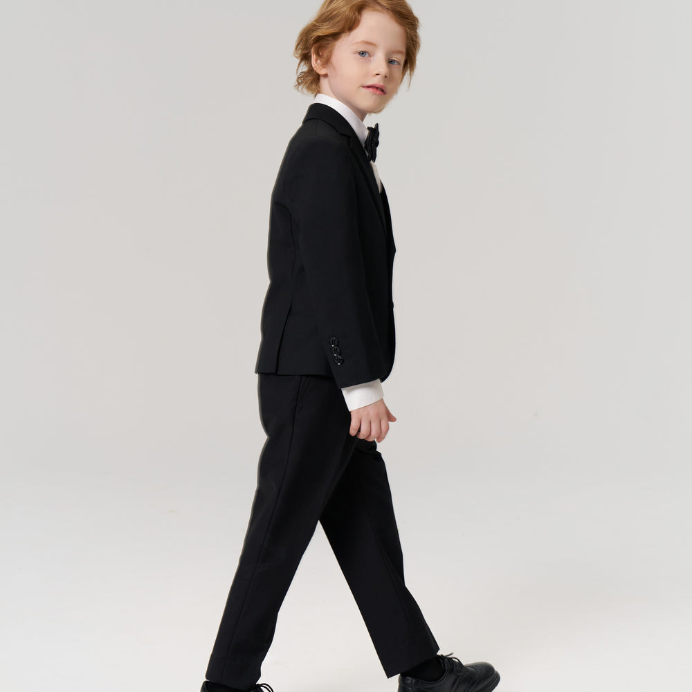 A young boy in a black suit, white shirt, and bow tie walks to the side against a plain light background.