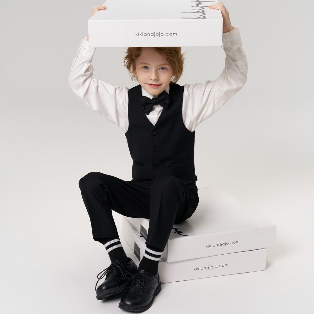 A young boy in a suit sits on stacked white boxes and holds another box over his head, all labeled "kikiandjojo.com," against a plain light background.