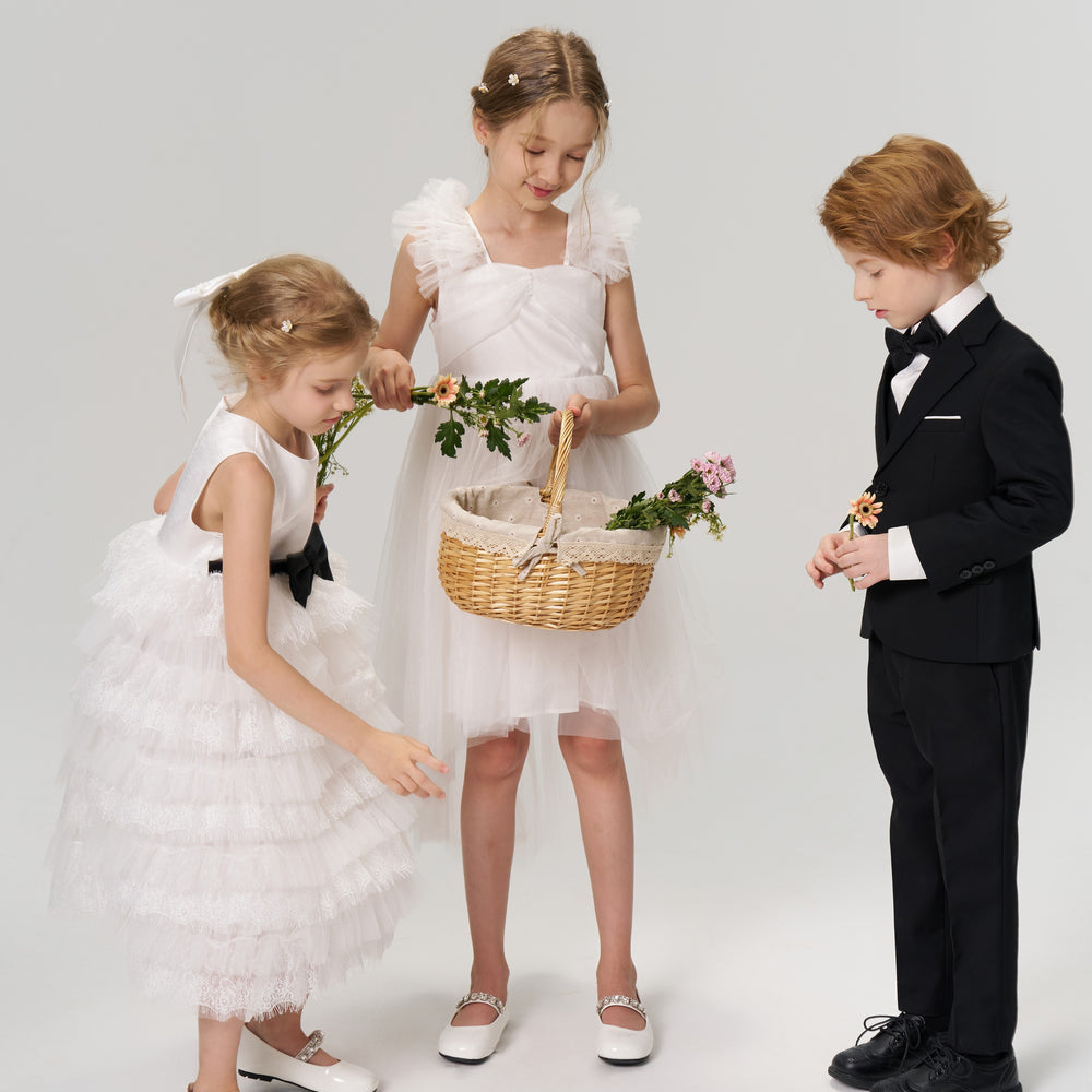 Three children dressed formally, two girls in white dresses with flowers and a basket, and a boy in a black suit, stand together against a plain background.