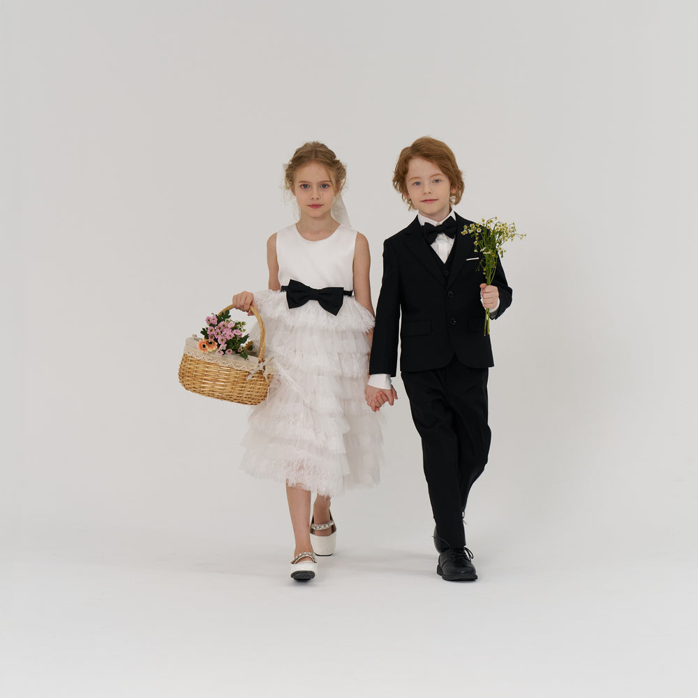 A young girl in a white dress holds a basket of flowers and walks hand-in-hand with a boy in a black suit who is holding a small bouquet, both facing forward against a plain background.
