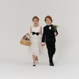 A young girl in a white dress holds a basket of flowers and walks hand-in-hand with a boy in a black suit who is holding a small bouquet, both facing forward against a plain background.