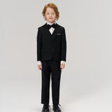 A young boy with light hair faces forward in KIKI and JOJO's Noir Bloom Suit Pants, styled with a black suit jacket, bow tie, and dress shoes—an ideal choice for formal occasions—set against a plain white background.