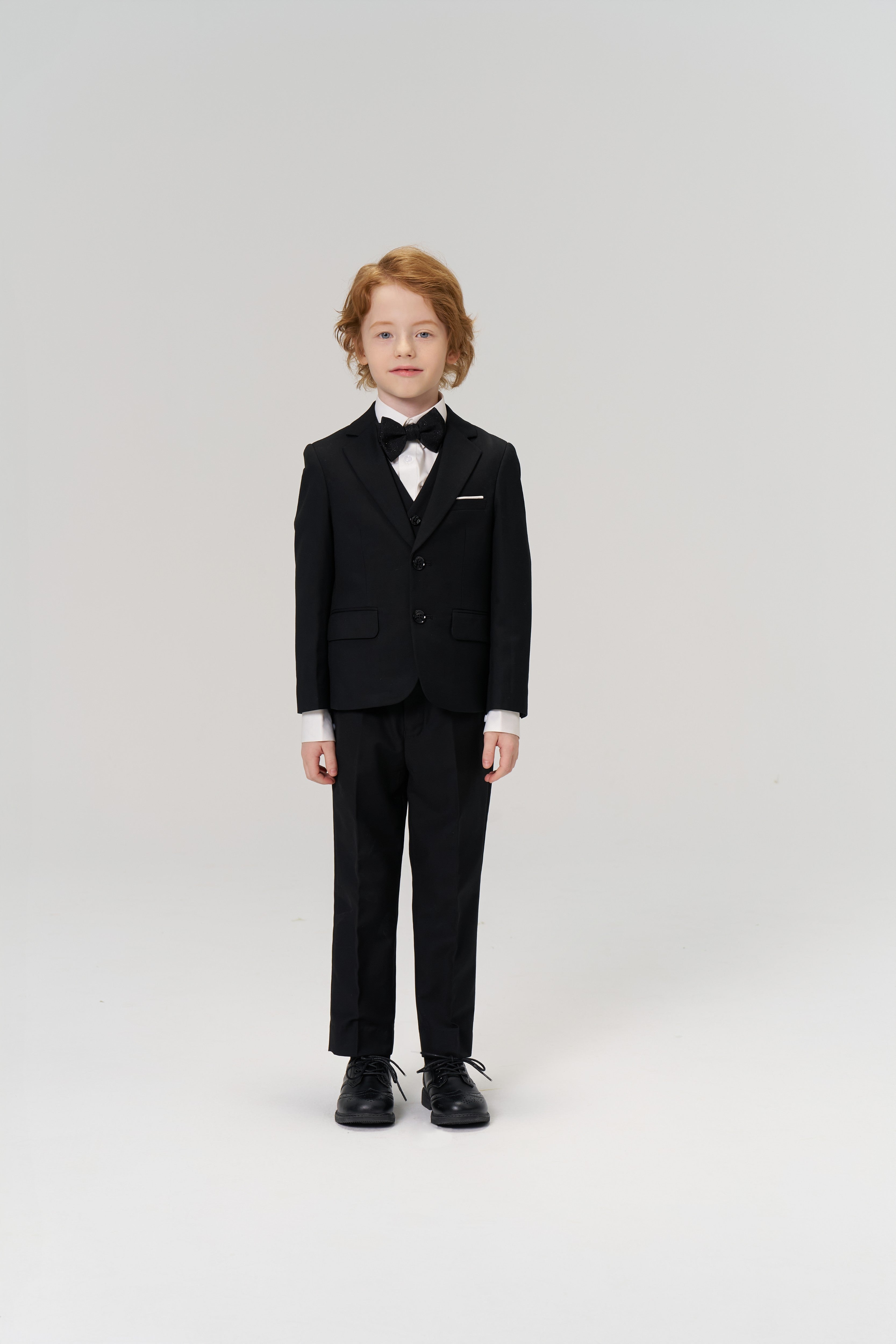 A young boy with light hair faces forward in KIKI and JOJO's Noir Bloom Suit Pants, styled with a black suit jacket, bow tie, and dress shoes—an ideal choice for formal occasions—set against a plain white background.